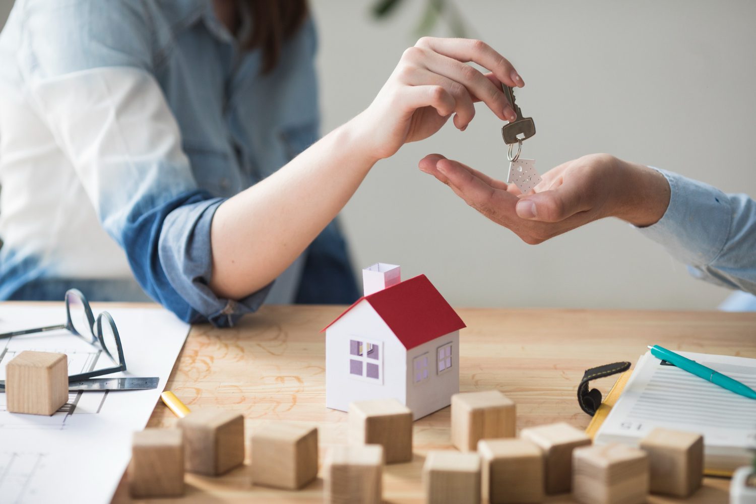 Close-Up Of A Woman Handing House Keys To A Man With A Miniature House Model On A Wooden Table